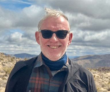 Smiling man with gray hair and sunglasses, standing outdoors in a desert landscape