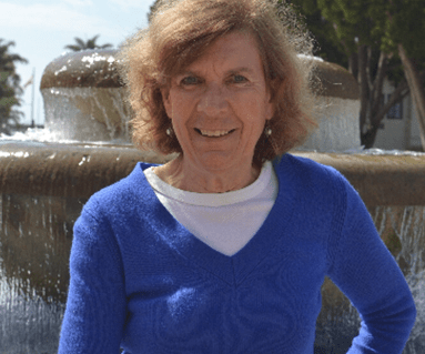 Smiling woman in a blue sweater stands by a fountain in a sunlit outdoor setting