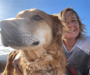 A woman smiles beside a golden dog in a bright landscape
