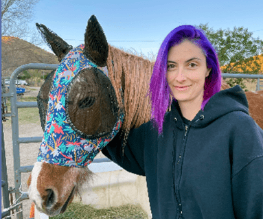 Woman with purple hair stands beside a horse wearing a floral-patterned fly mask