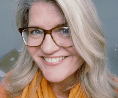 A smiling woman with long blonde hair wearing glasses and an orange scarf