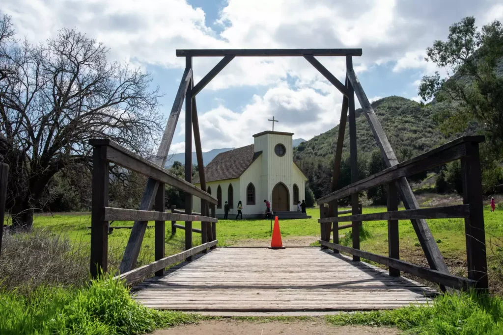 A quaint church stands beyond a wooden bridge, surrounded by greenery and mountains, with visitors exploring the serene landscape.